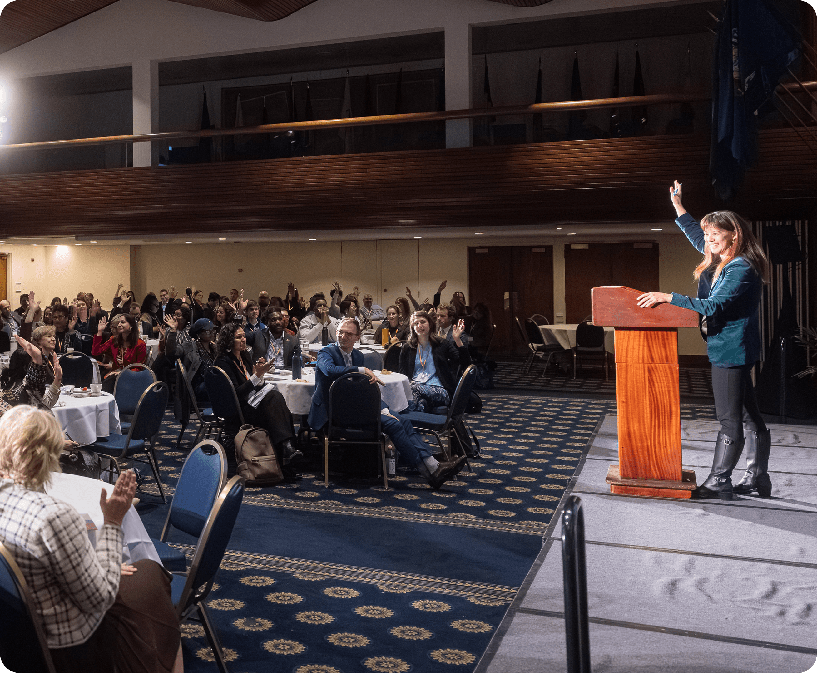 Members participate in a Coalition convening in a large hall, with a speaker at a podium and attendees at round tables with hands raised